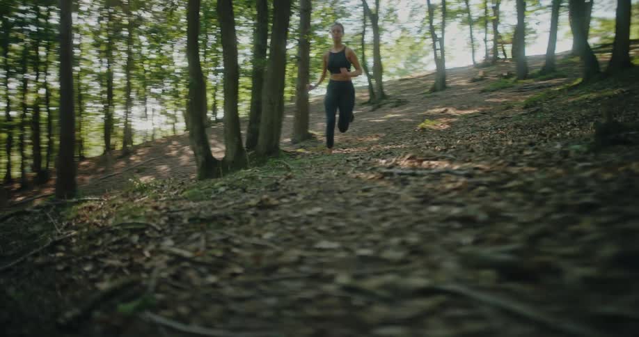 An athletic woman trail runs downhill through a sunlit forest with the camera tracking her feet