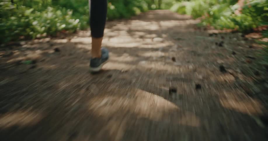 A low-angle tracking shot follows a runner's feet as they pound a dirt trail through a lush, sunlit forest