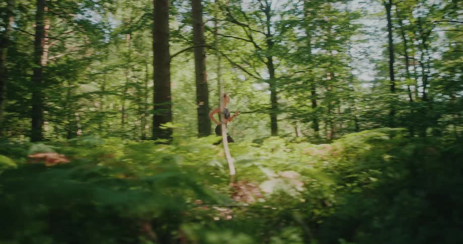 Slow motion side view of an athletic woman jogging through a sun-dappled forest with lush green ferns