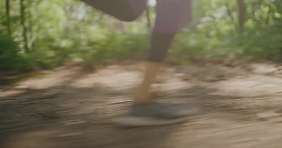 A low-angle close-up of a person's running shoes as they jog along a dirt path through a sunny forest