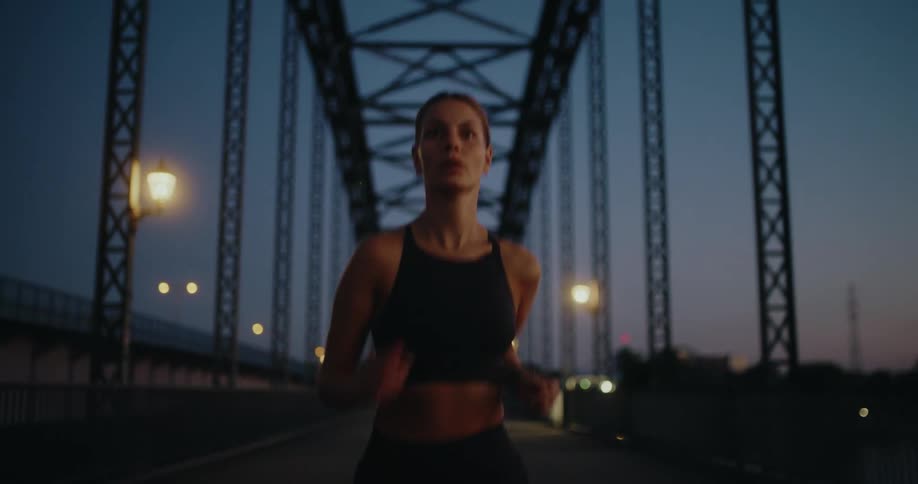 A fit young woman in a sports bra runs with determination across a steel bridge in the early morning twilight
