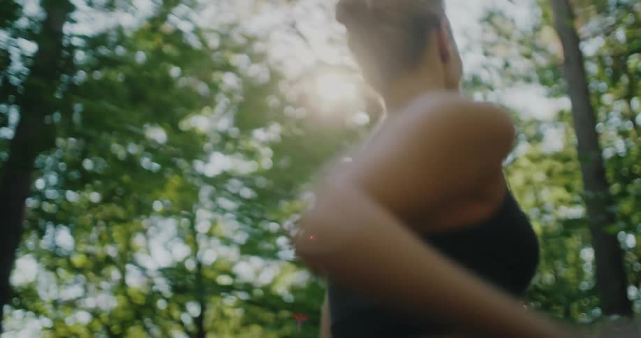 Low-angle follow shot of a woman jogging through a sunlit forest trail in slow motion