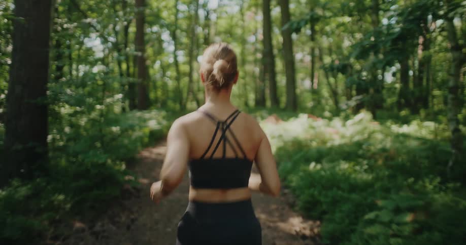 Back view of a fit woman in athletic wear running along a scenic, sun-dappled trail in a lush green forest