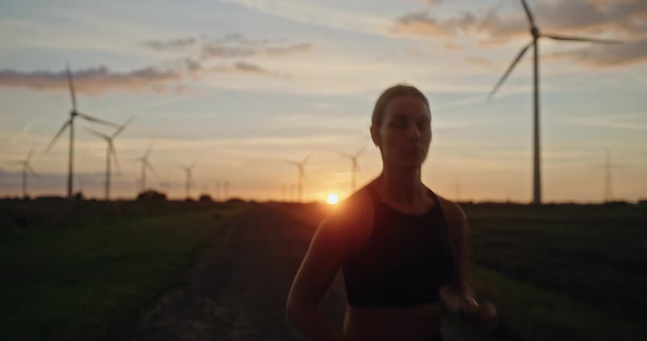 A determined woman runs on a rural road through a wind farm at sunset in slow motion