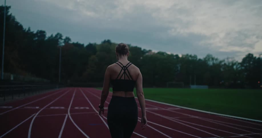 A determined female athlete walks along an empty running track in the early morning or late evening, preparing for her run