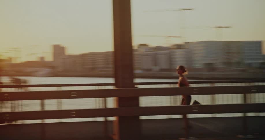 Side view of a woman in sportswear running across a bridge with an industrial city skyline in the background at sunset