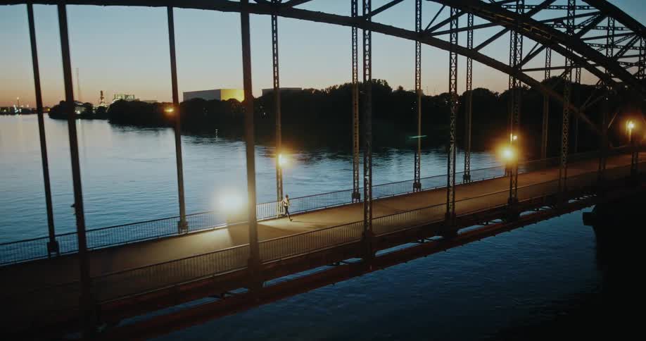 An aerial view follows a lone runner jogging across an illuminated steel arch bridge over a wide river at dusk