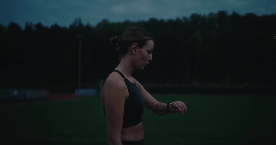A determined woman in athletic wear checks her smartwatch on a sports field at dusk before walking out of frame