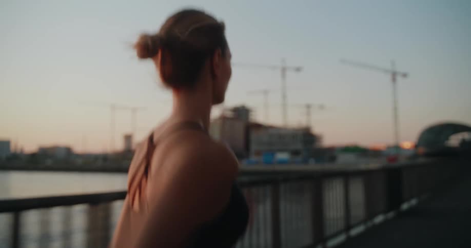 Slow motion shot from behind of a woman jogging across an industrial waterfront bridge at sunset
