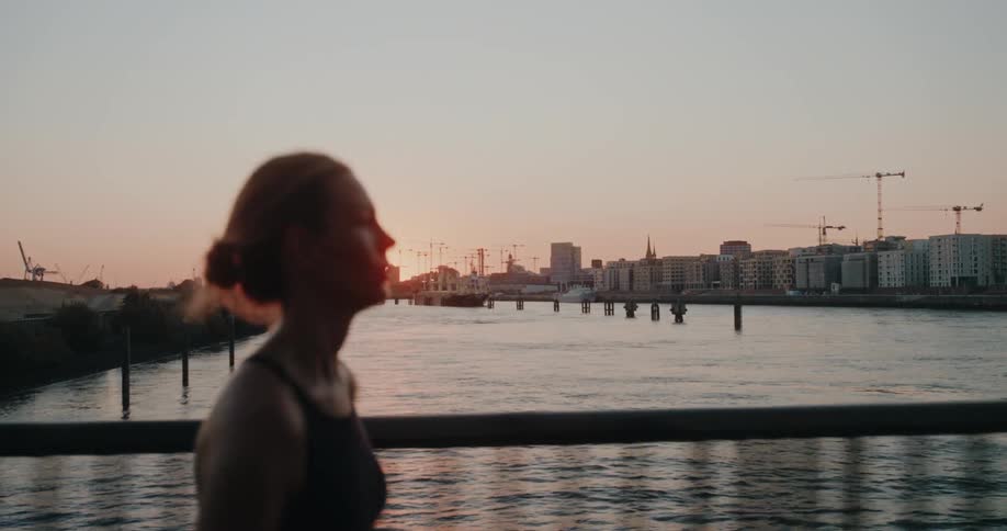 Cinematic slow-motion shot of a woman jogging along a city riverfront during a beautiful sunset