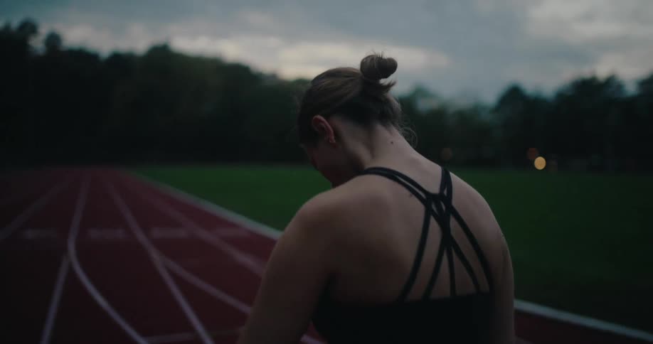 A determined female runner ties her hair into a bun on an empty track at twilight, focused and ready to train