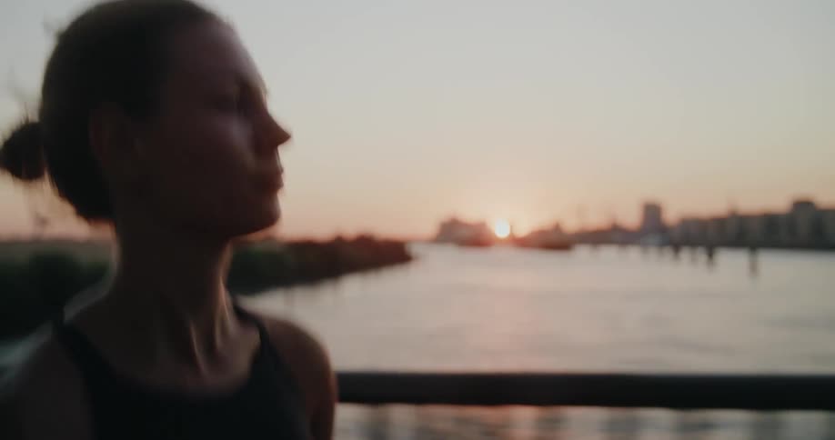 Slow motion side view of an athletic woman jogging on a bridge with the city skyline and a golden sunset in the background