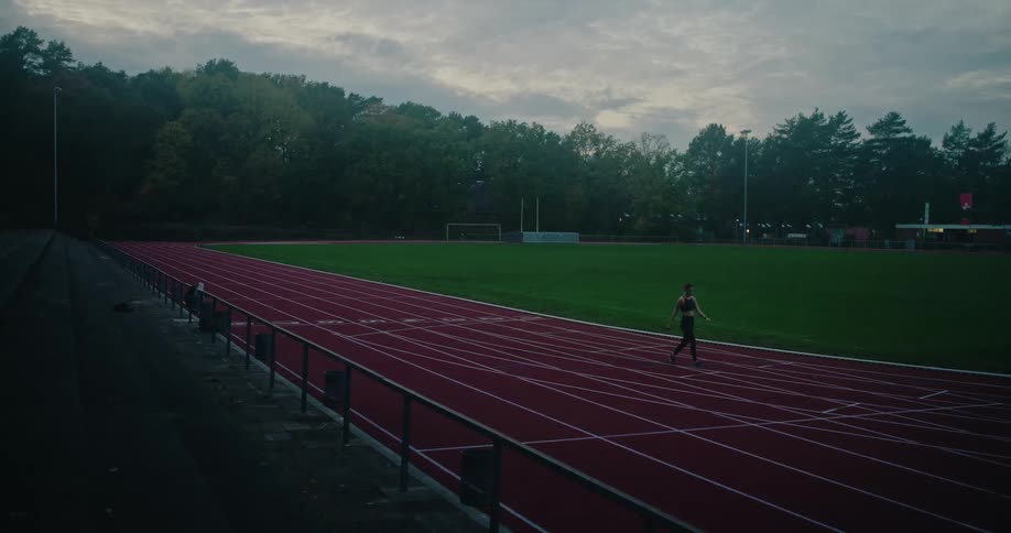 A fit woman runs towards the camera before slowing to a walk on an empty athletic track at dusk