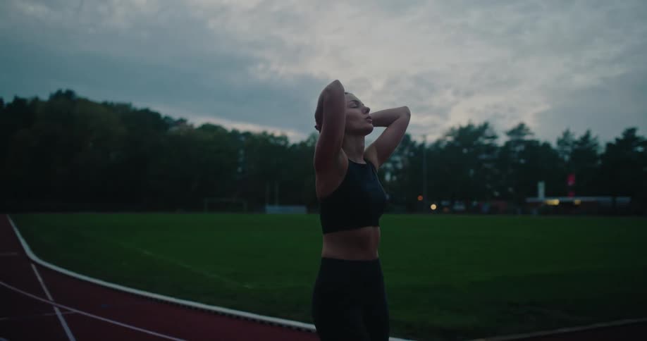A focused female athlete walks and stretches on a running track during a moody evening workout