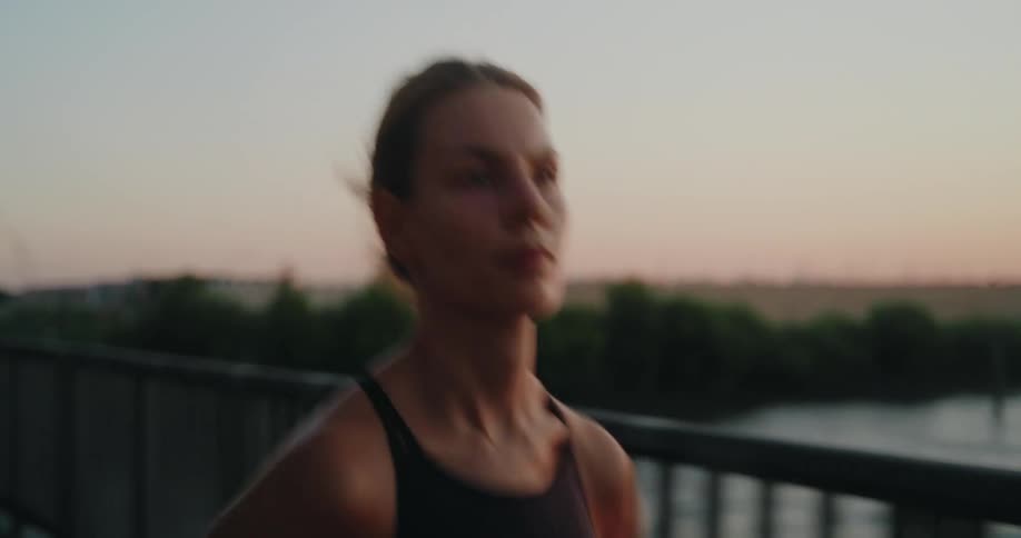 Slow motion close-up of a determined woman in athletic wear jogging across a bridge during a beautiful, warm sunset