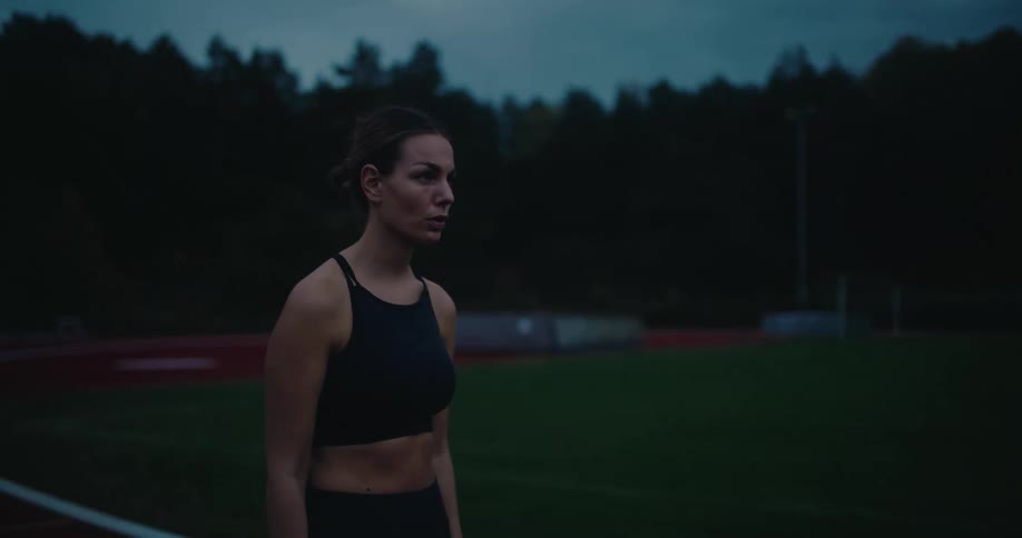 A determined female athlete in a black sports bra checks her watch before training on an outdoor track at dusk