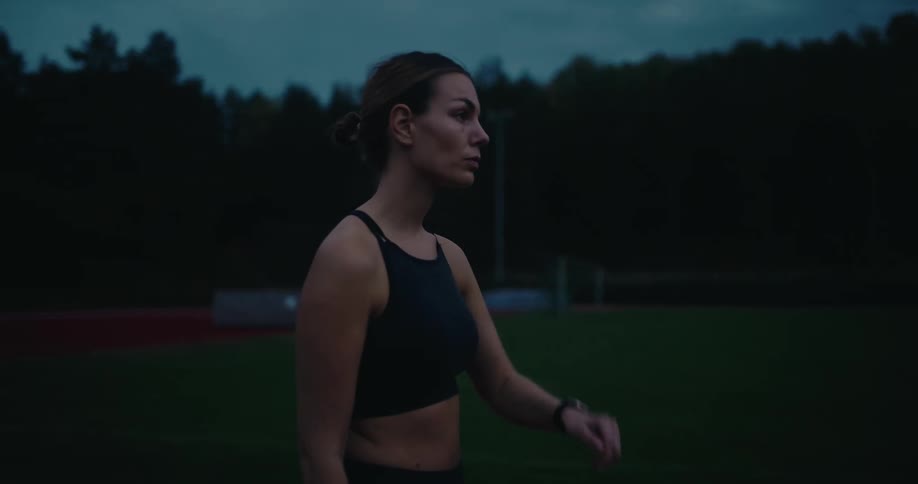 A determined female athlete checks her smartwatch and prepares for an intense workout on a running track at dusk