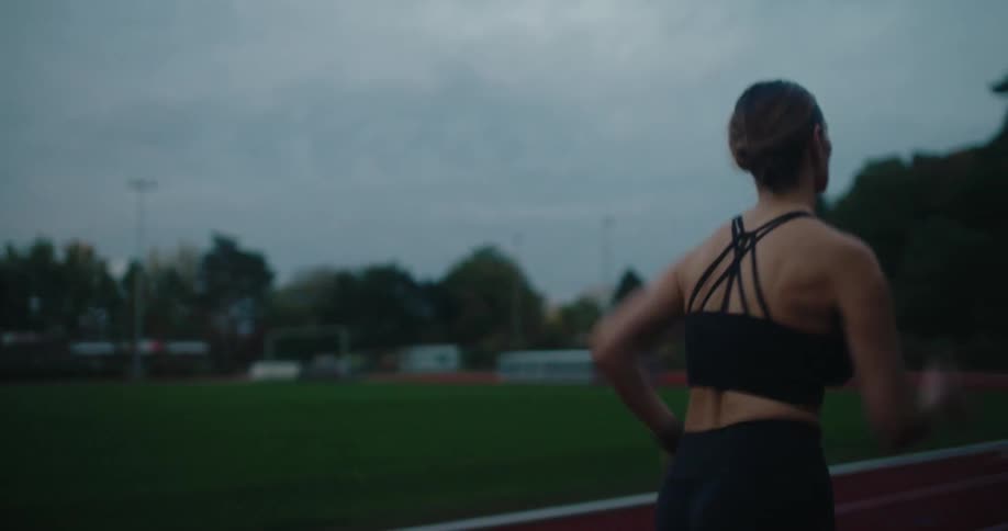 A determined female runner trains on an athletic track at dusk, slowing down to check her watch