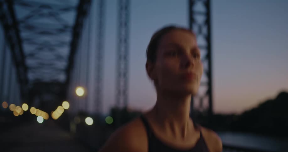 A focused athletic woman runs across an urban bridge at dusk, with the city lights creating a beautiful bokeh in the background