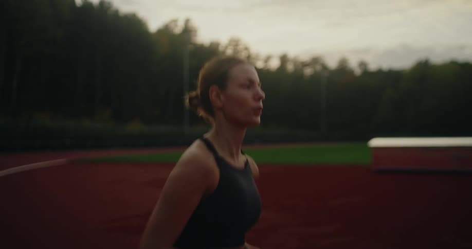 A focused female runner trains on a red athletic track at a stadium during dusk