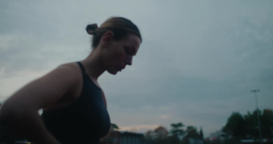 An athletic woman finishes her run on an outdoor track at dusk, stopping to catch her breath and recover