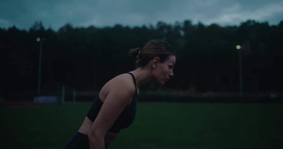 A determined female athlete prepares to sprint in slow motion on a dark outdoor track at dusk