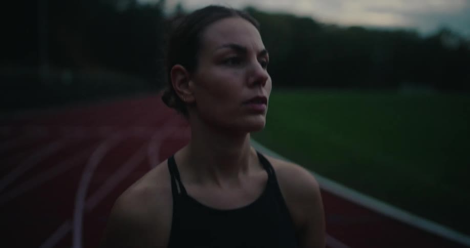 A determined female athlete takes a moment to catch her breath and refocus on a running track during a moody evening workout