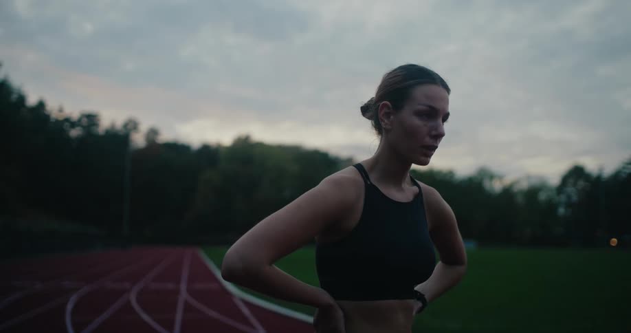 A tired female runner catches her breath and looks up at the sky while resting on a running track at dusk