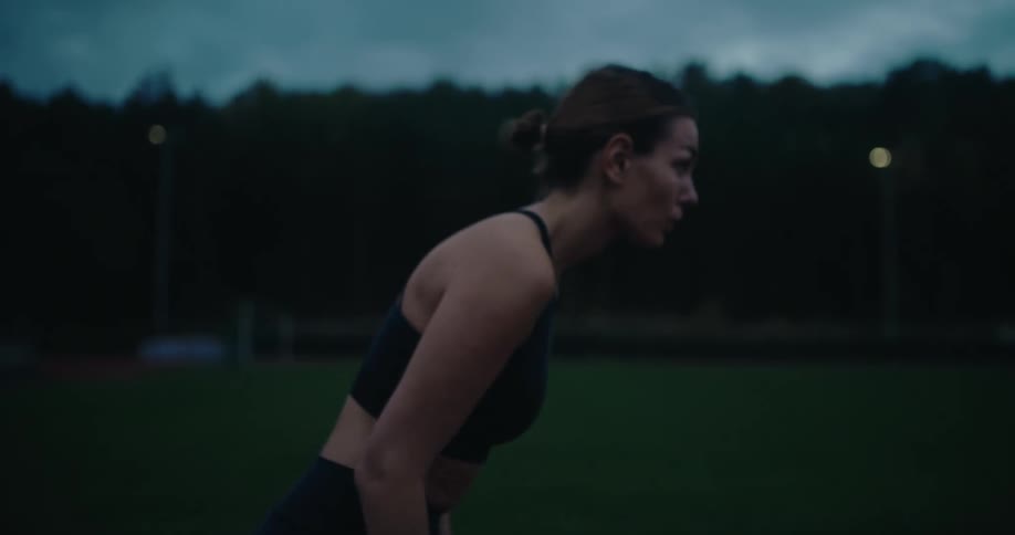 In slow motion, a determined female runner catches her breath, gets into starting position, and sprints off on an outdoor track at dusk