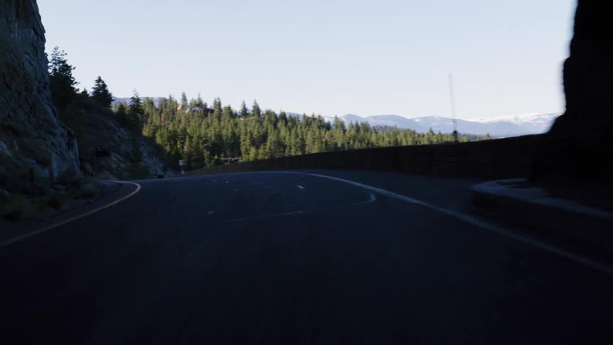 A point-of-view shot driving slowly out of a dark mountain tunnel onto a scenic, winding road surrounded by pine forests