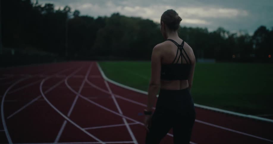 In slow motion, a focused female athlete ties her hair up and warms up on a running track at dusk before her training session