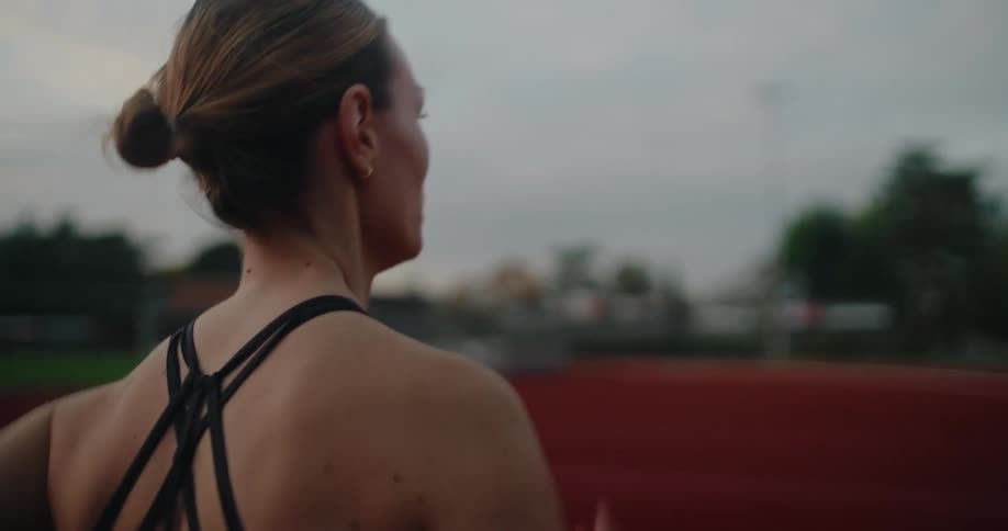 A determined athletic woman in a sports bra runs along a red track at dusk in a focused slow motion workout