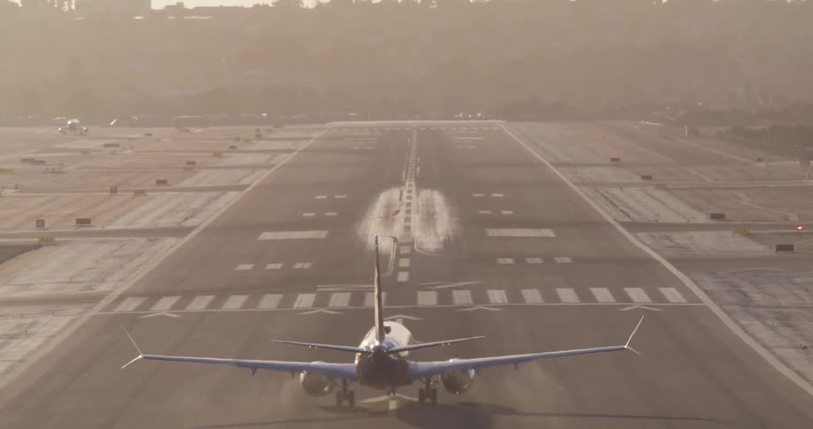 Rear view of a commercial airliner taking off from an airport runway in slow motion against the hazy morning sun