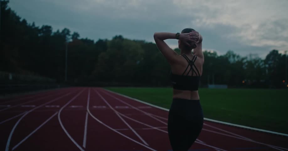 A determined female athlete stretches and prepares for a run on an empty track at dusk