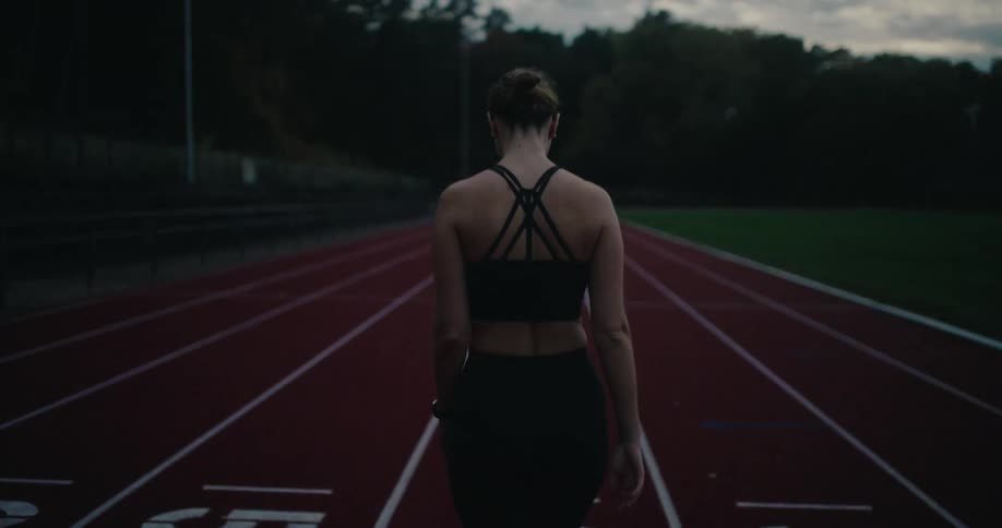 A woman in athletic wear stretches and walks down a red running track at dusk in slow motion