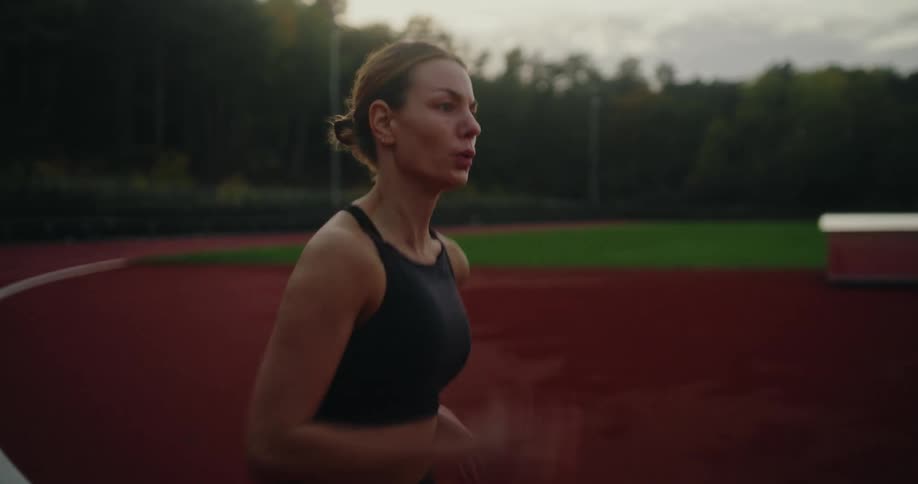 A focused and determined woman in black sportswear runs in slow motion on a red track at an outdoor stadium