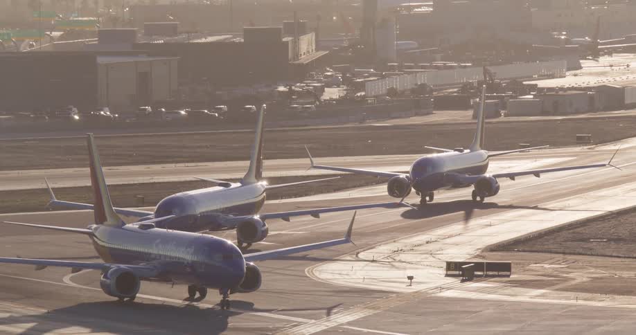 A lineup of three Southwest Airlines passenger jets waits on the tarmac at a busy airport during a hazy golden hour