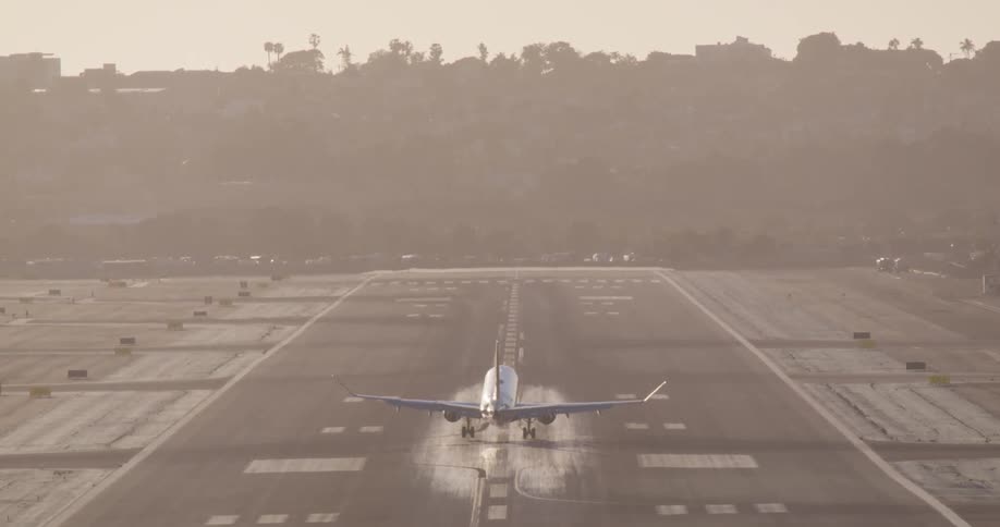 A slow motion, telephoto shot captures the rear view of a commercial airliner as it descends and lands on a hazy runway at sunset, with smoke puffing from the tires on touchdown