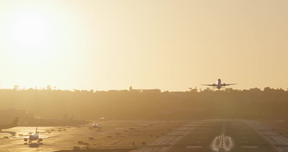 A passenger airplane takes off from the runway in slow motion against the hazy golden glow of the setting sun