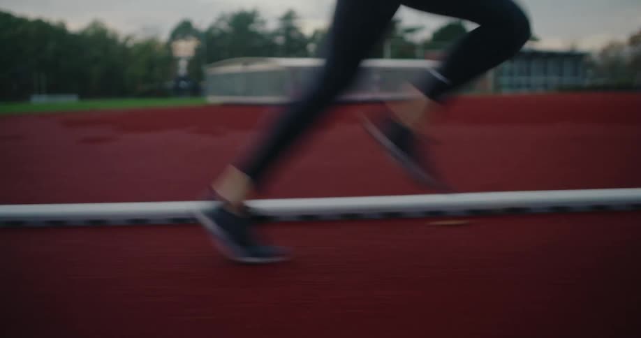 A low-angle tracking shot captures the powerful motion of a runner's legs sprinting along a red athletic track