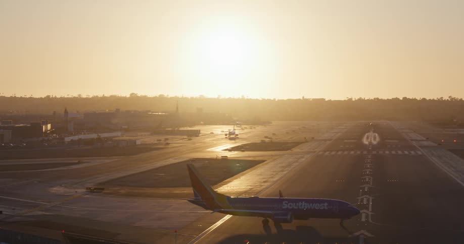 A wide shot shows several airplanes taxiing and landing on the runway of a busy airport during a hazy golden sunset