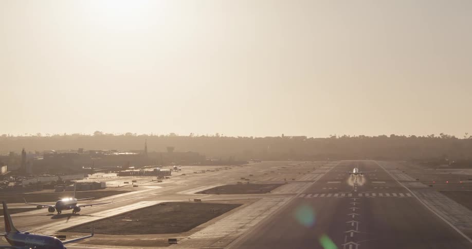 A commercial airplane takes off from a busy airport runway, ascending into the hazy sky during a golden sunset