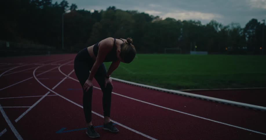 A fit woman in athletic wear catches her breath on a running track after a strenuous workout at dusk