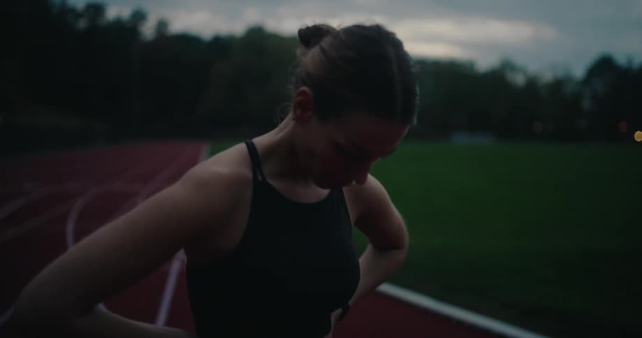 A determined female athlete catches her breath on a running track after a workout at dusk before walking away