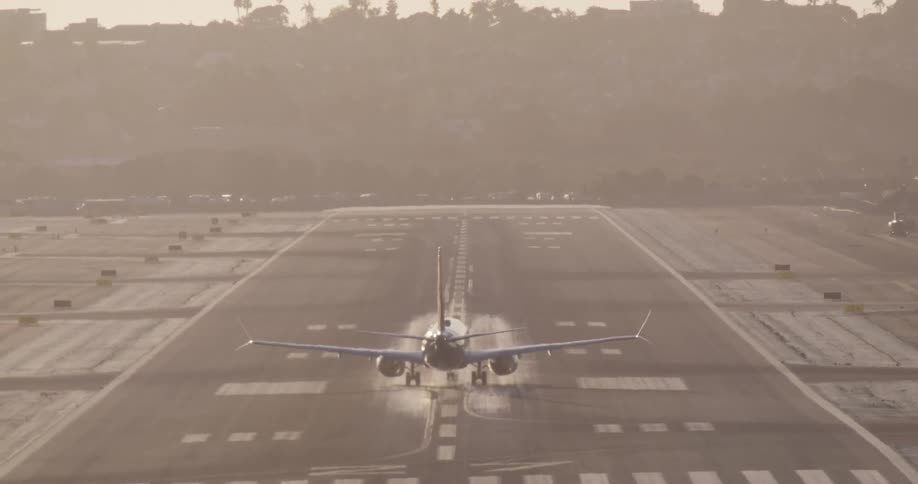 From behind, a commercial jet accelerates down a long runway and gracefully takes off into the hazy sky