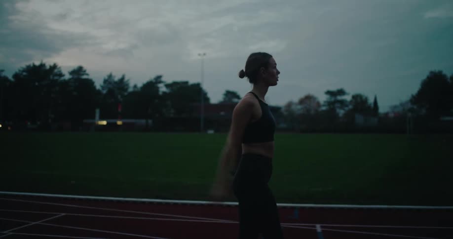A determined female athlete warms up on an outdoor running track at dusk before her training session
