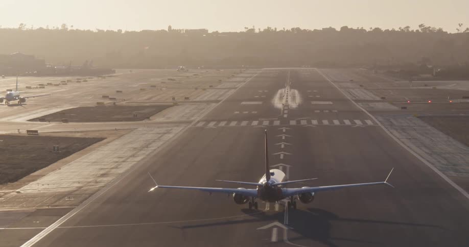 A passenger jet taxis down a long runway before turning around and taking off into the hazy sunrise