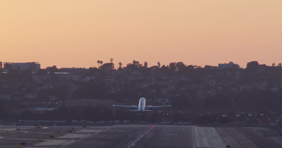 From a direct rear view, a passenger jet accelerates down the runway and takes off into the warm, hazy sky at dawn