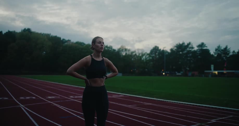 A focused female runner in black sportswear completes her workout on a track, slowing down to check her smartwatch before stretching and catching her breath under a cloudy evening sky