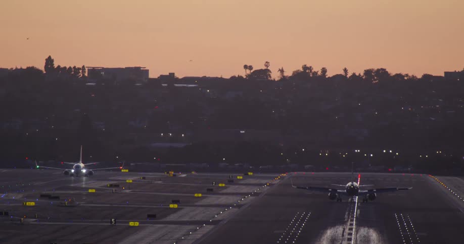 Two commercial airplanes with lights flashing taxi on the runways of a busy airport during a warm, picturesque sunset